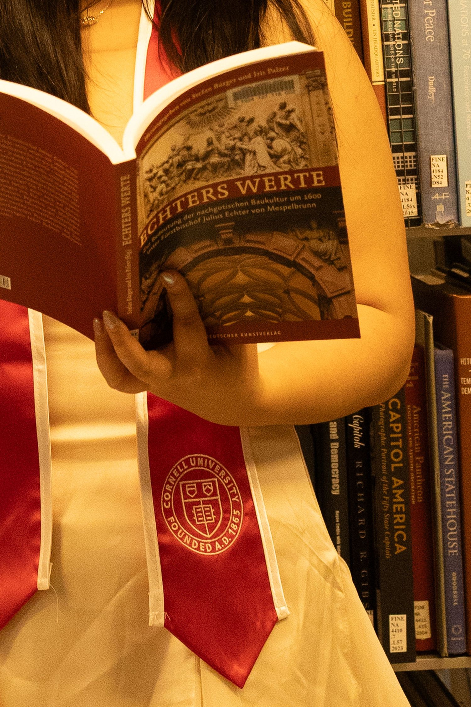 Cornell University graduate in red sash reads German book “Richters Werte” in a library filled with academic texts.
