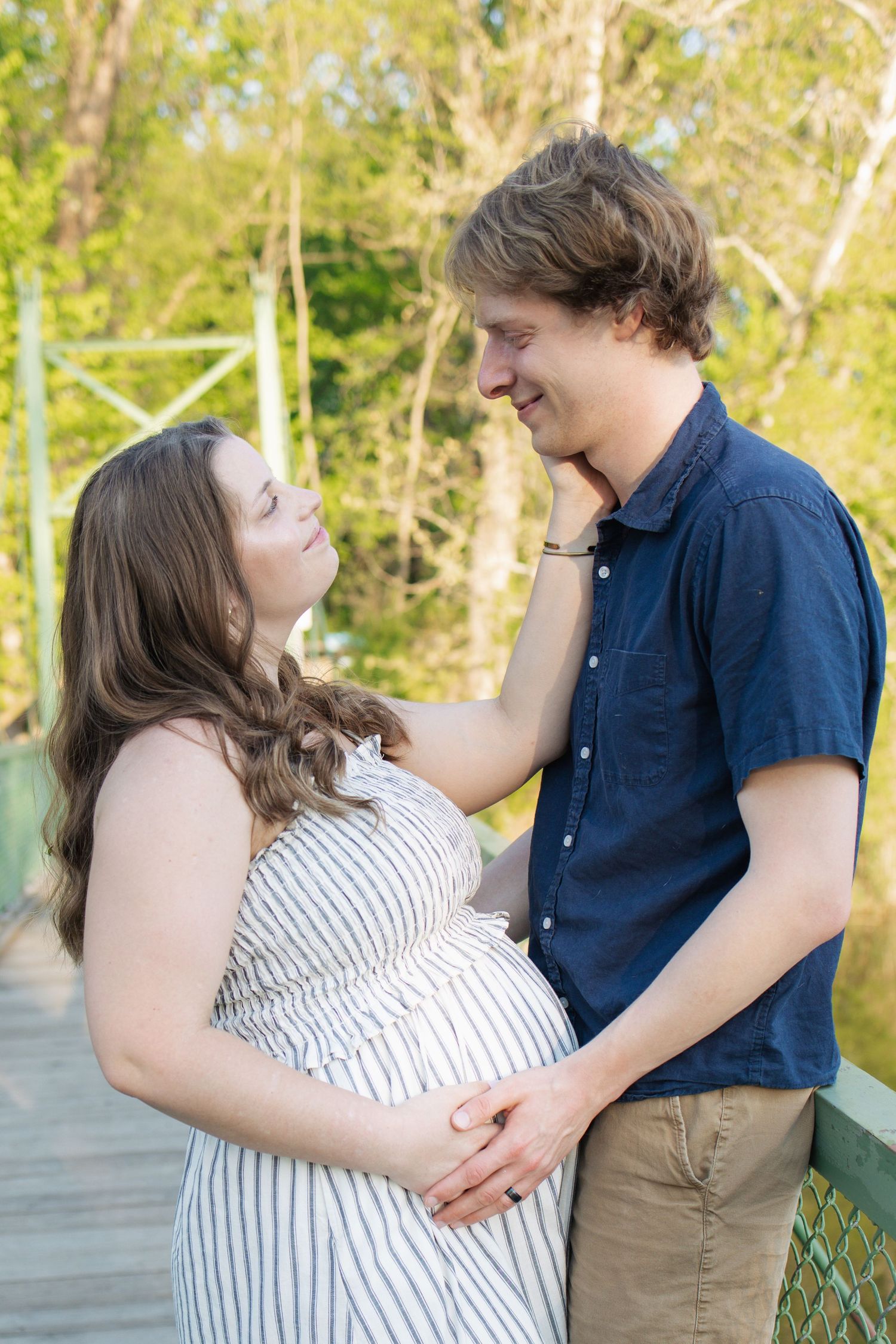 Happy couple sharing a tender moment outdoors, pregnant woman in striped dress with partner on a bridge surrounded by trees.