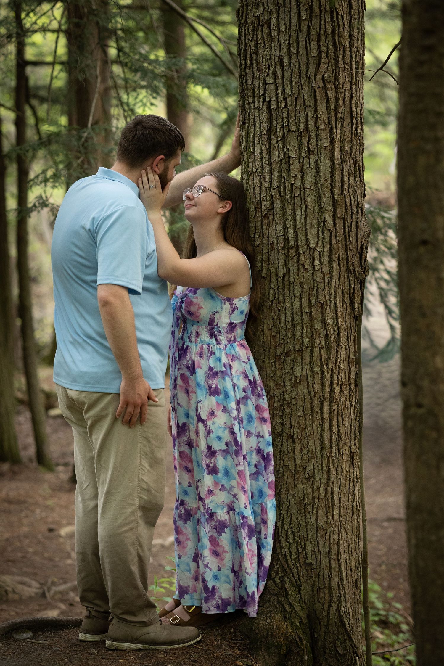 Couple sharing a romantic moment leaning against a tree in a forest, woman in floral maxi dress smiling at man.