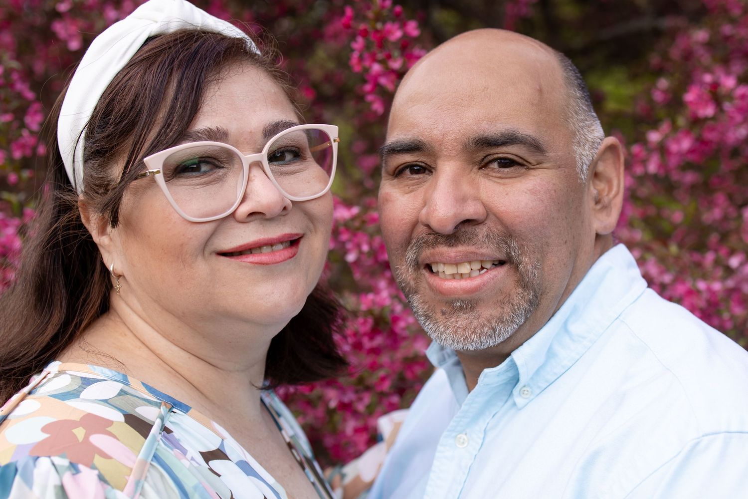 Smiling couple posing together outdoors with pink flowering trees in background, woman wearing glasses and floral top.