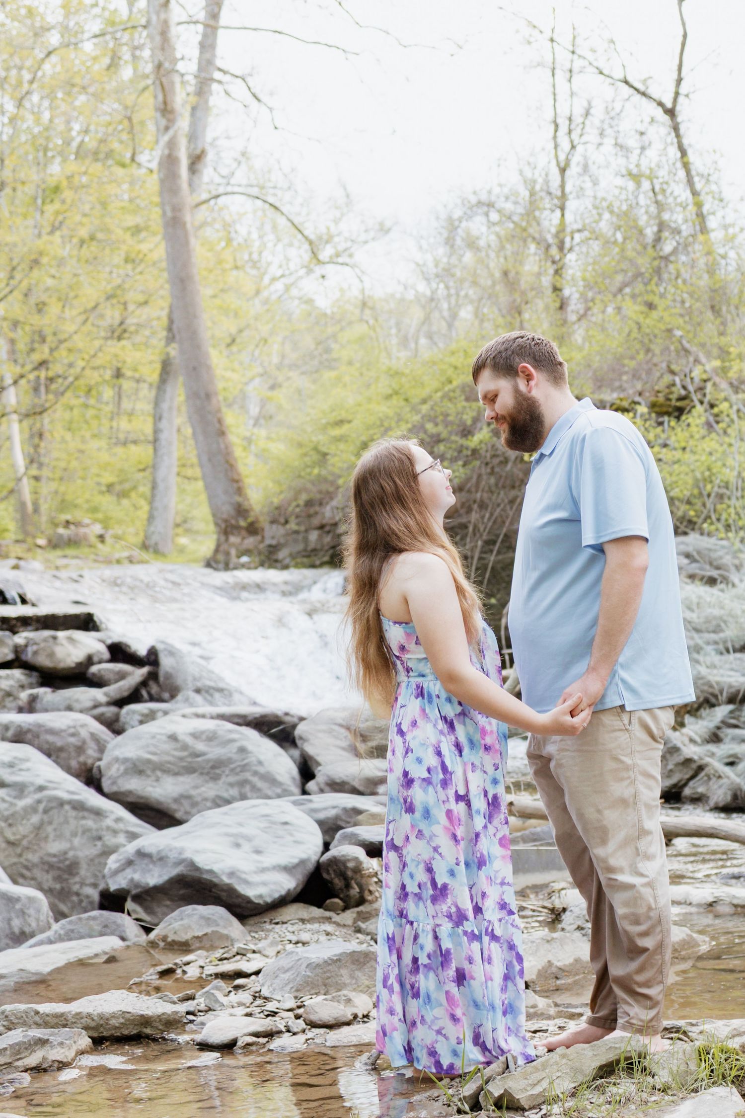 Couple holding hands by a rocky waterfall, woman in floral maxi dress, man in blue shirt, surrounded by spring trees.