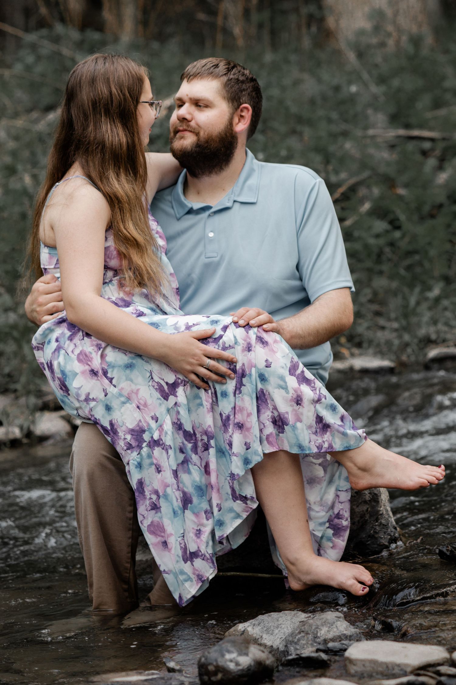 Couple sitting on rocks in a creek, woman in floral dress on man's lap, sharing a tender moment outdoors.