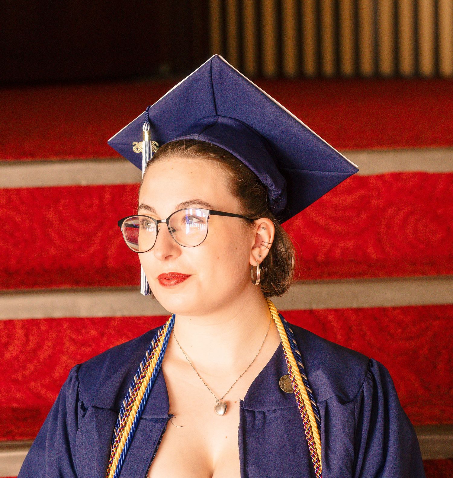Female graduate in blue cap and gown with glasses and red lipstick poses on red carpeted stairs.