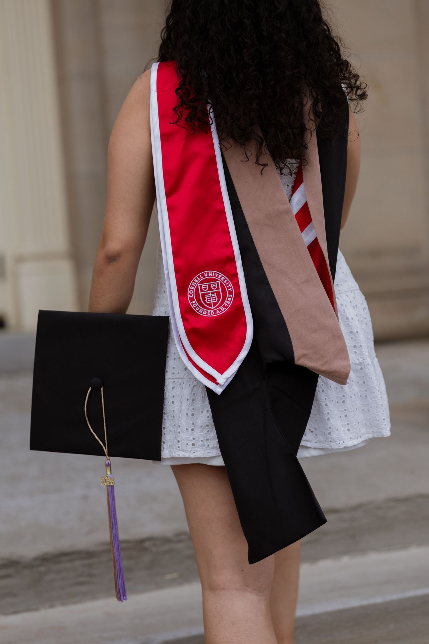 Graduate holding black cap with purple tassel, wearing red university stole over white dress at graduation ceremony.