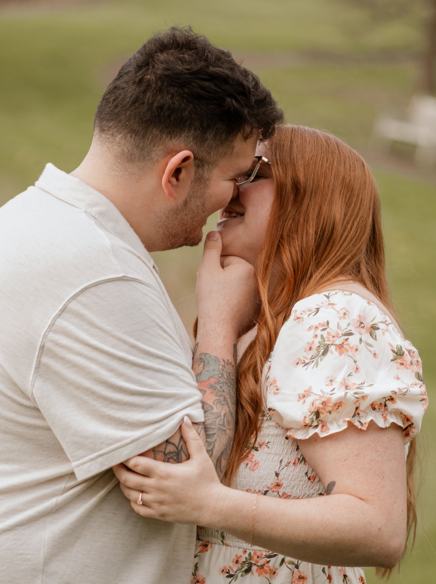Romantic couple nearly kissing outdoors, woman with long red hair in floral dress, man with tattoos.