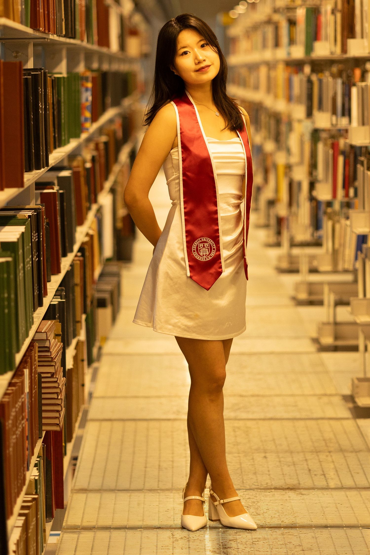 Young woman in white dress and red graduation sash poses between library bookshelves in white heels.