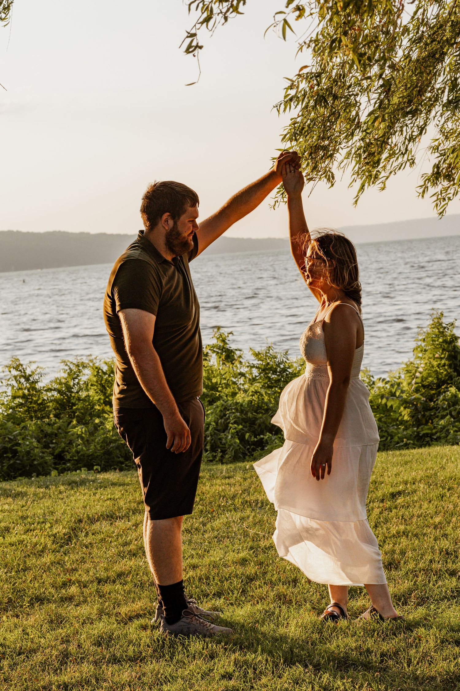 Couple dancing outdoors by a lake at golden hour, man spinning woman in white dress on green grass.