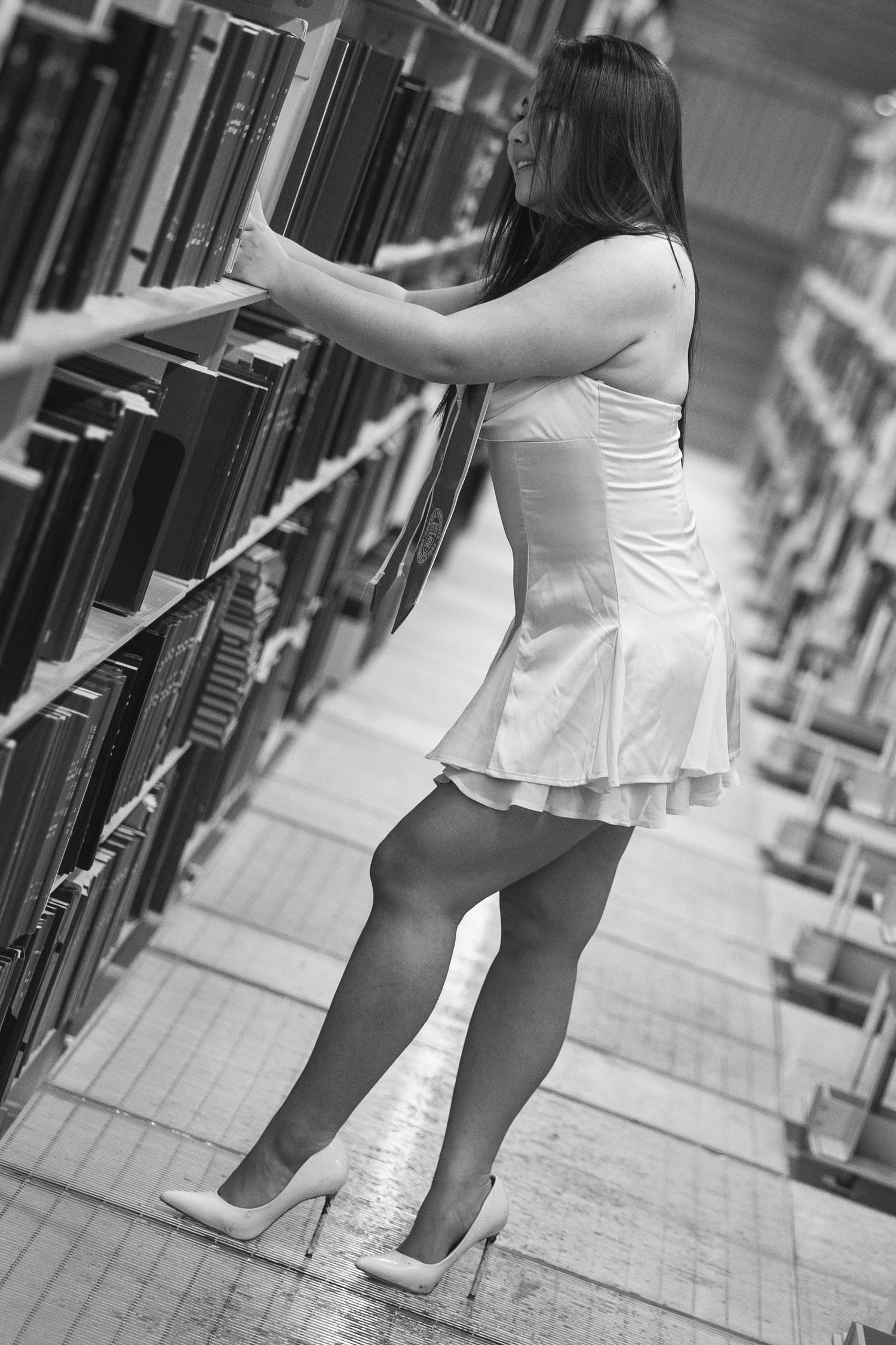 Black and white photo of a woman in a white dress and heels browsing books on shelves in a library.