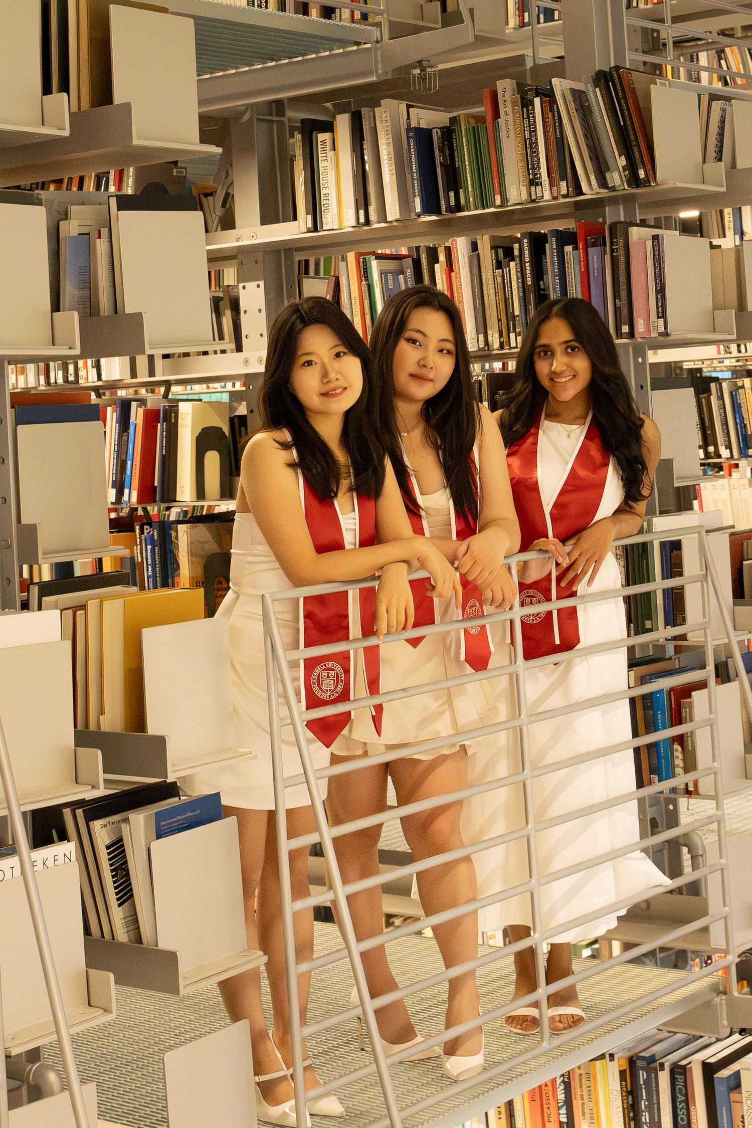 Three young women in white dresses and red graduation sashes pose proudly in a library with bookshelves behind them.
