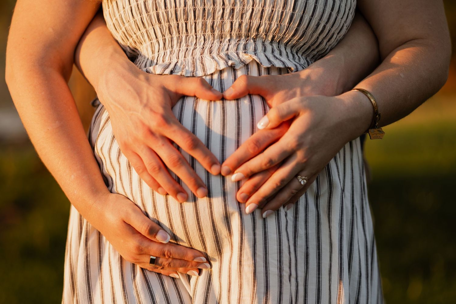 Pregnant woman in striped dress with two pairs of hands forming a heart shape on her baby bump in golden sunlight.