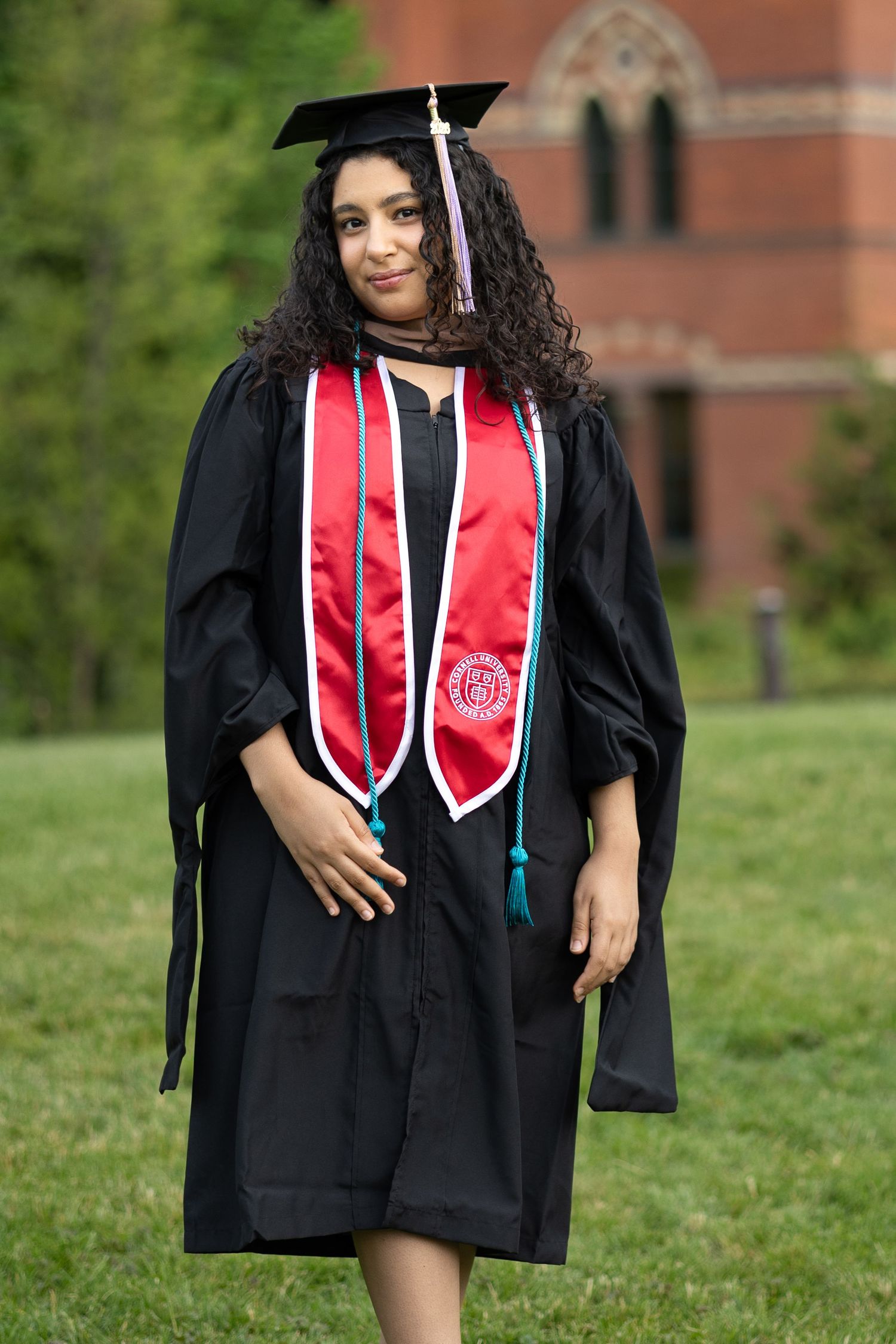 Young woman in black graduation cap and gown with red sash poses outdoors on green campus lawn.