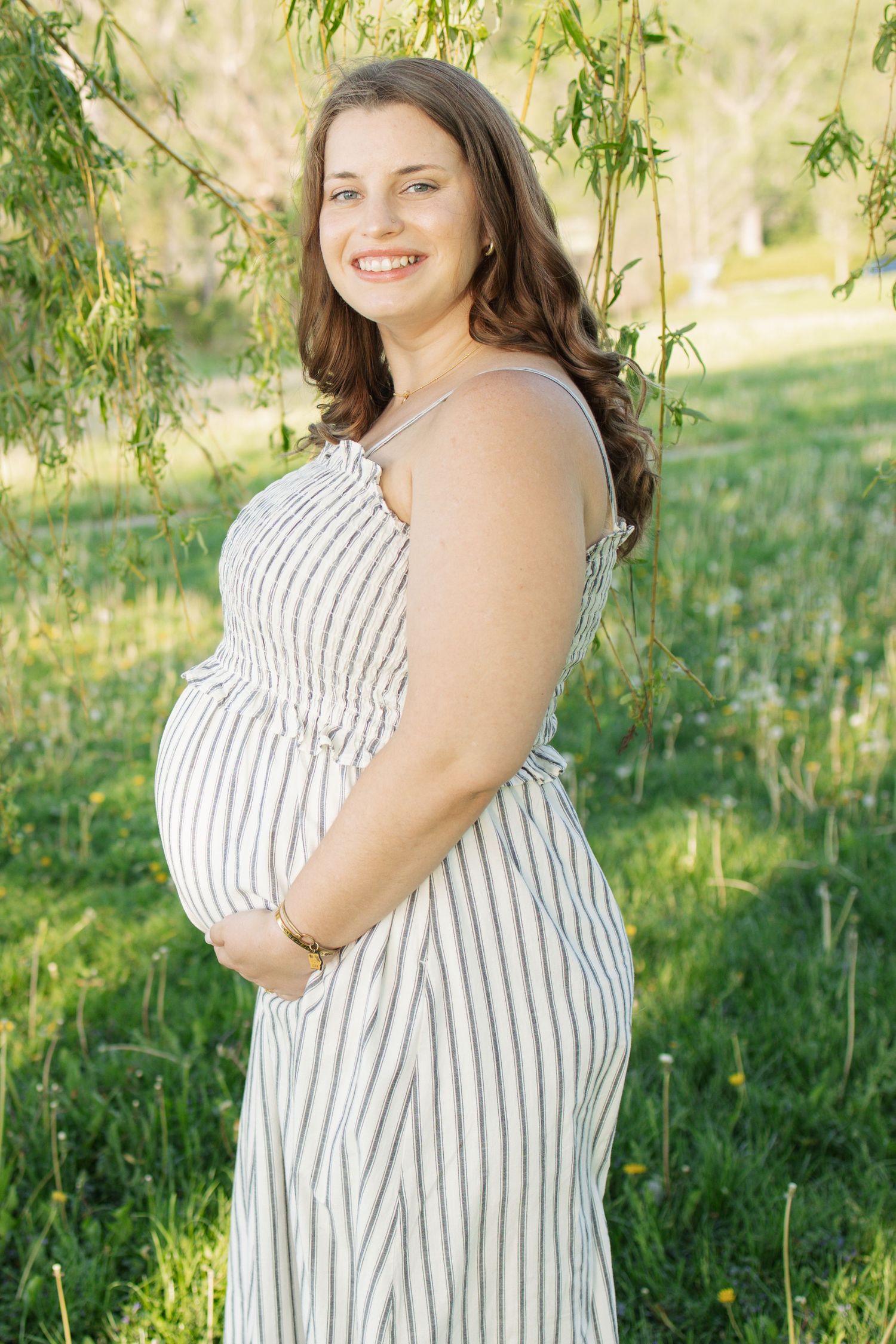 Smiling pregnant woman in striped dress holding belly outdoors near willow tree on sunny day.