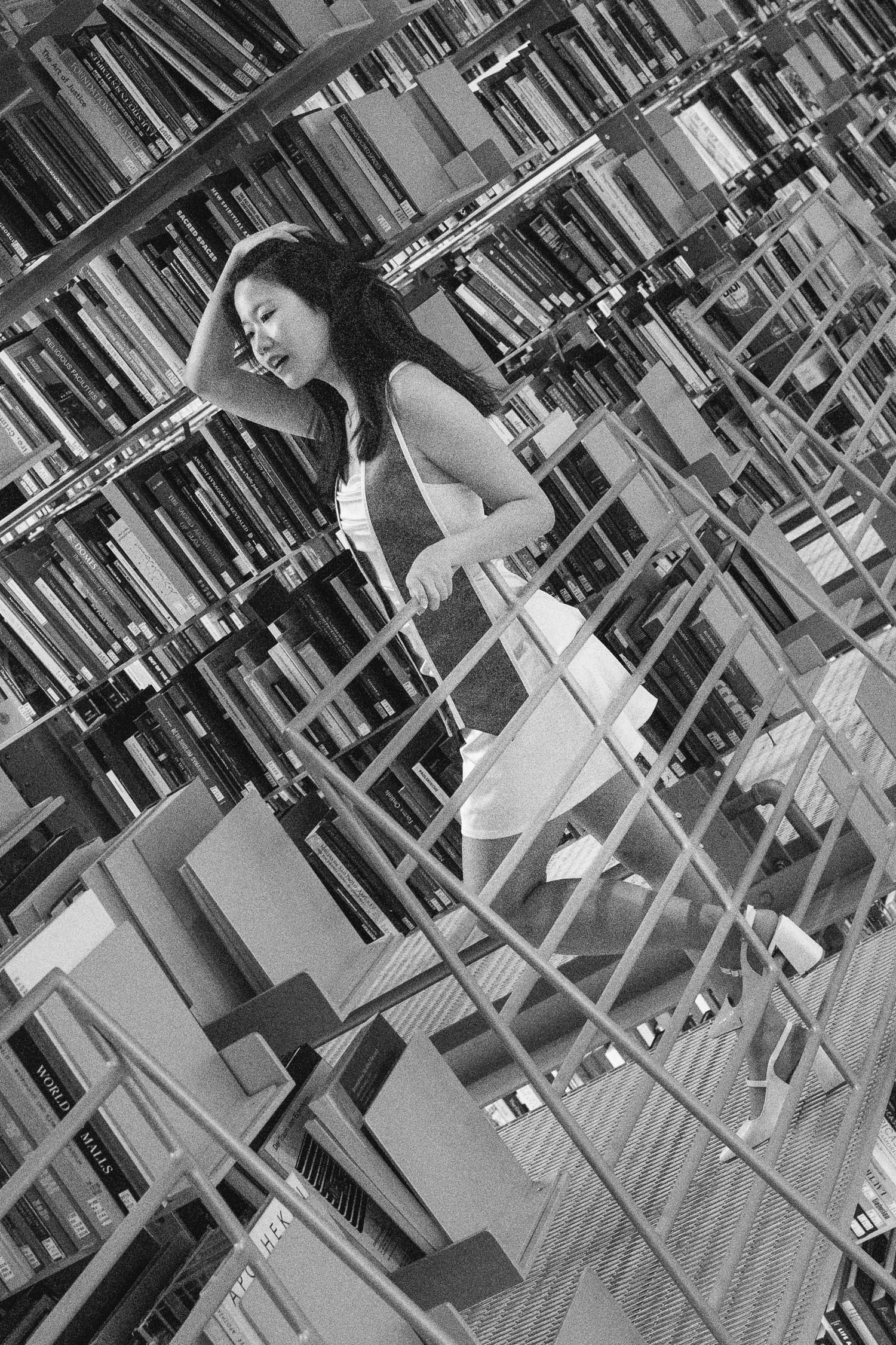 Black and white photo of a young woman browsing books in a multi-level library with tall shelves.