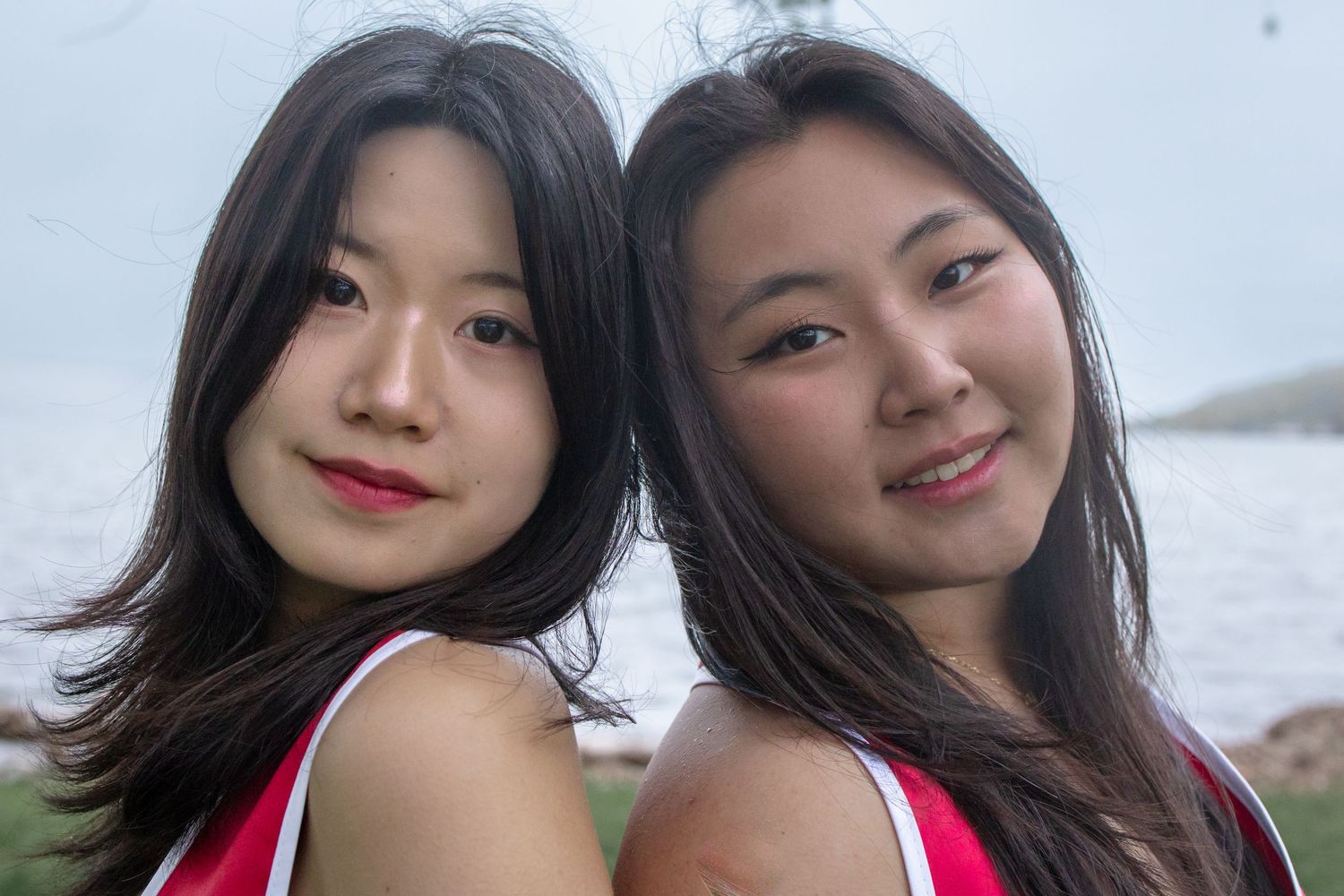 Two young Asian women smiling outdoors near water, wearing red tops, with long dark hair.