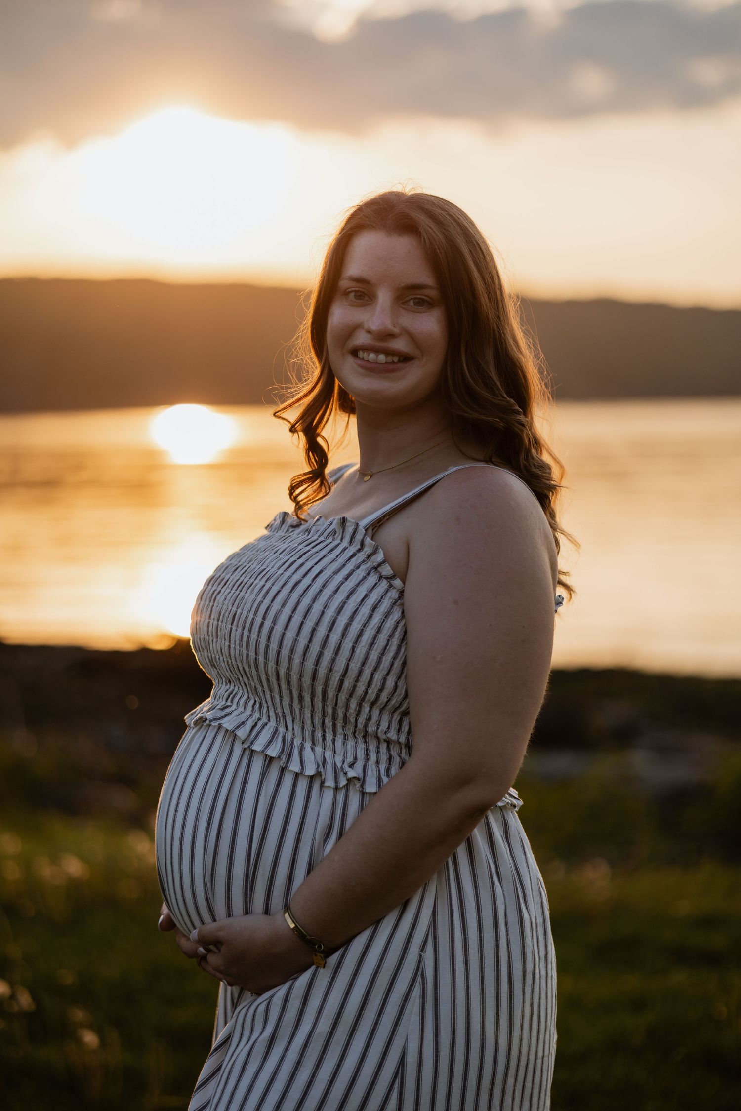 Smiling pregnant woman in striped dress holding baby bump at golden sunset by the water.