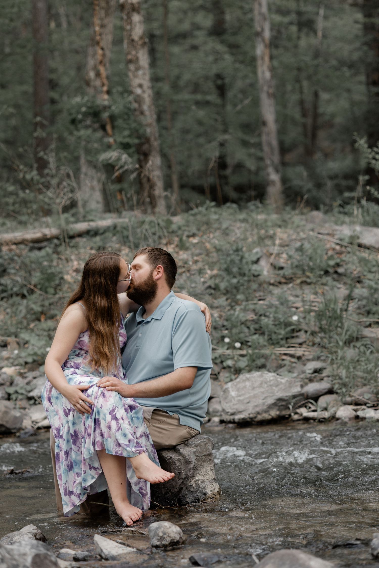 Couple sharing a kiss while sitting on rocks by a forest stream, woman in floral dress, man in blue shirt.