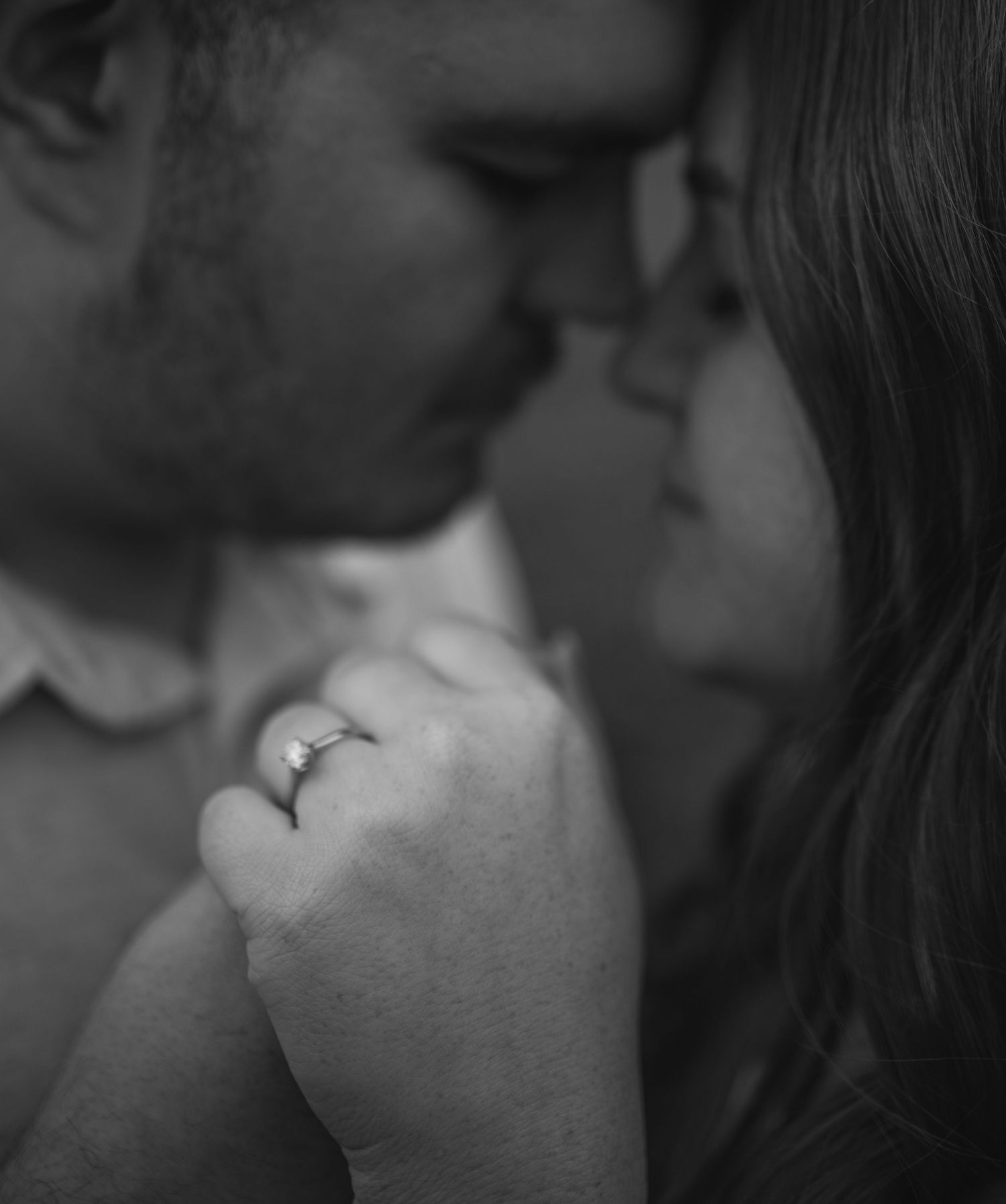 Black and white close-up of couple sharing intimate moment, woman displaying diamond engagement ring.