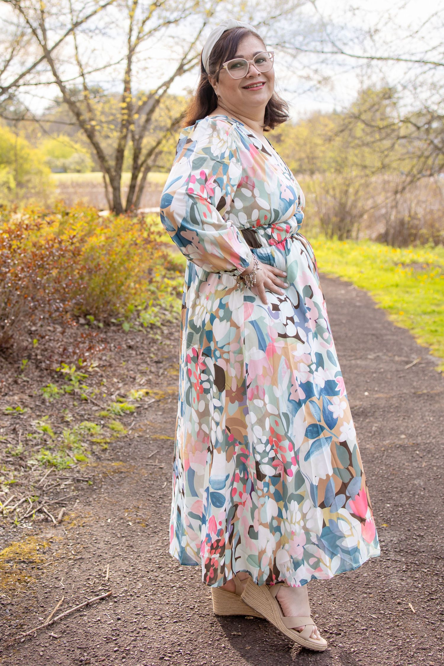 Woman in floral maxi dress with glasses poses on garden path surrounded by autumn foliage.