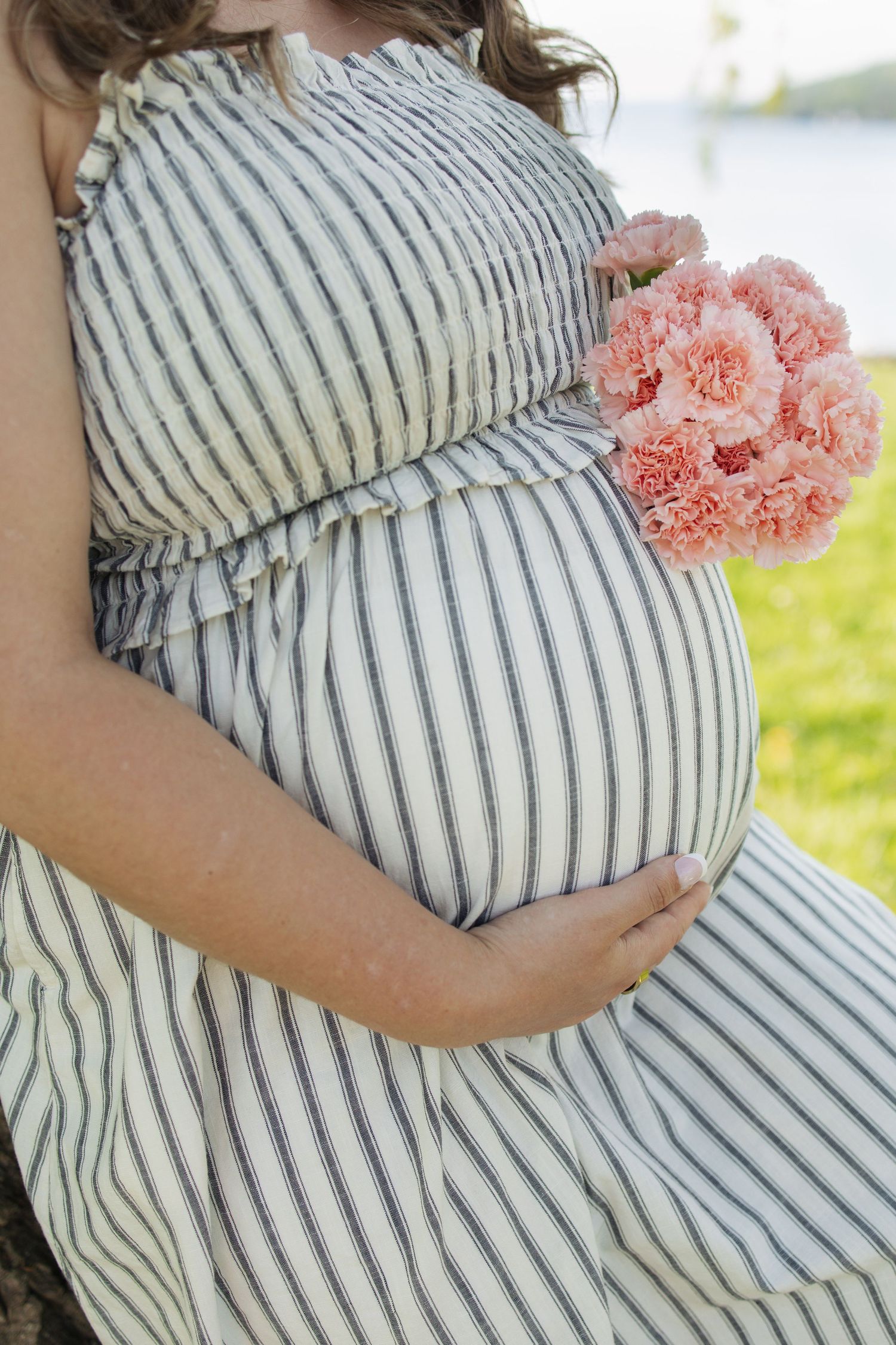 Pregnant woman in striped dress holding pink carnations while cradling her baby bump outdoors.