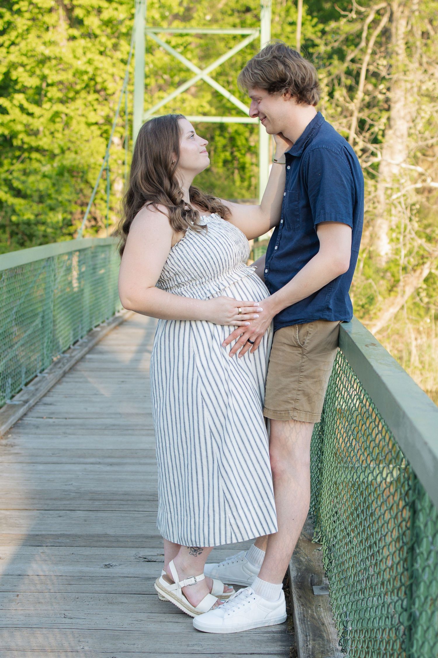 Couple on a bridge holding pregnant belly in striped dress and navy shirt outdoors.
