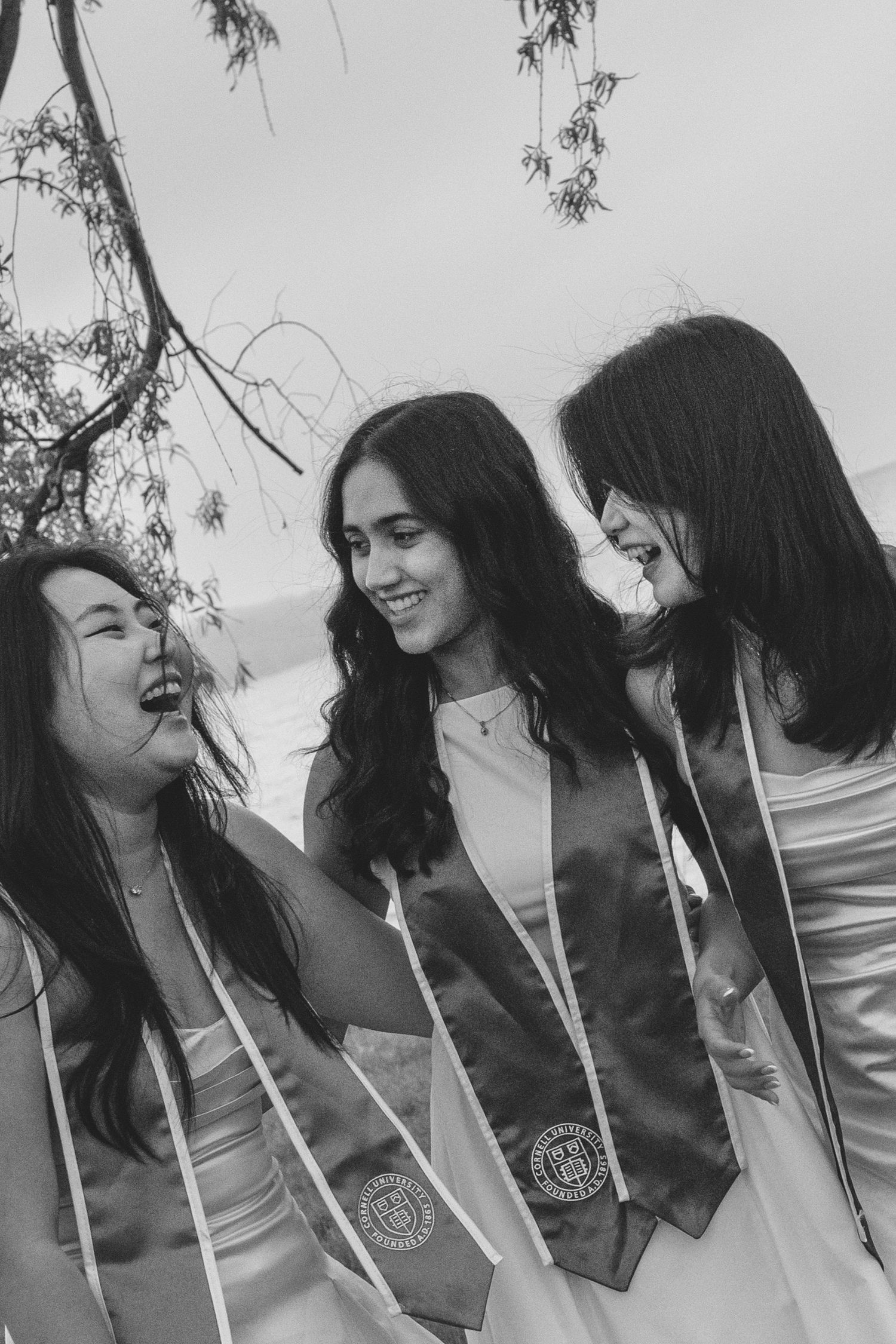 Three smiling women in graduation sashes celebrate outdoors in a black and white photo.