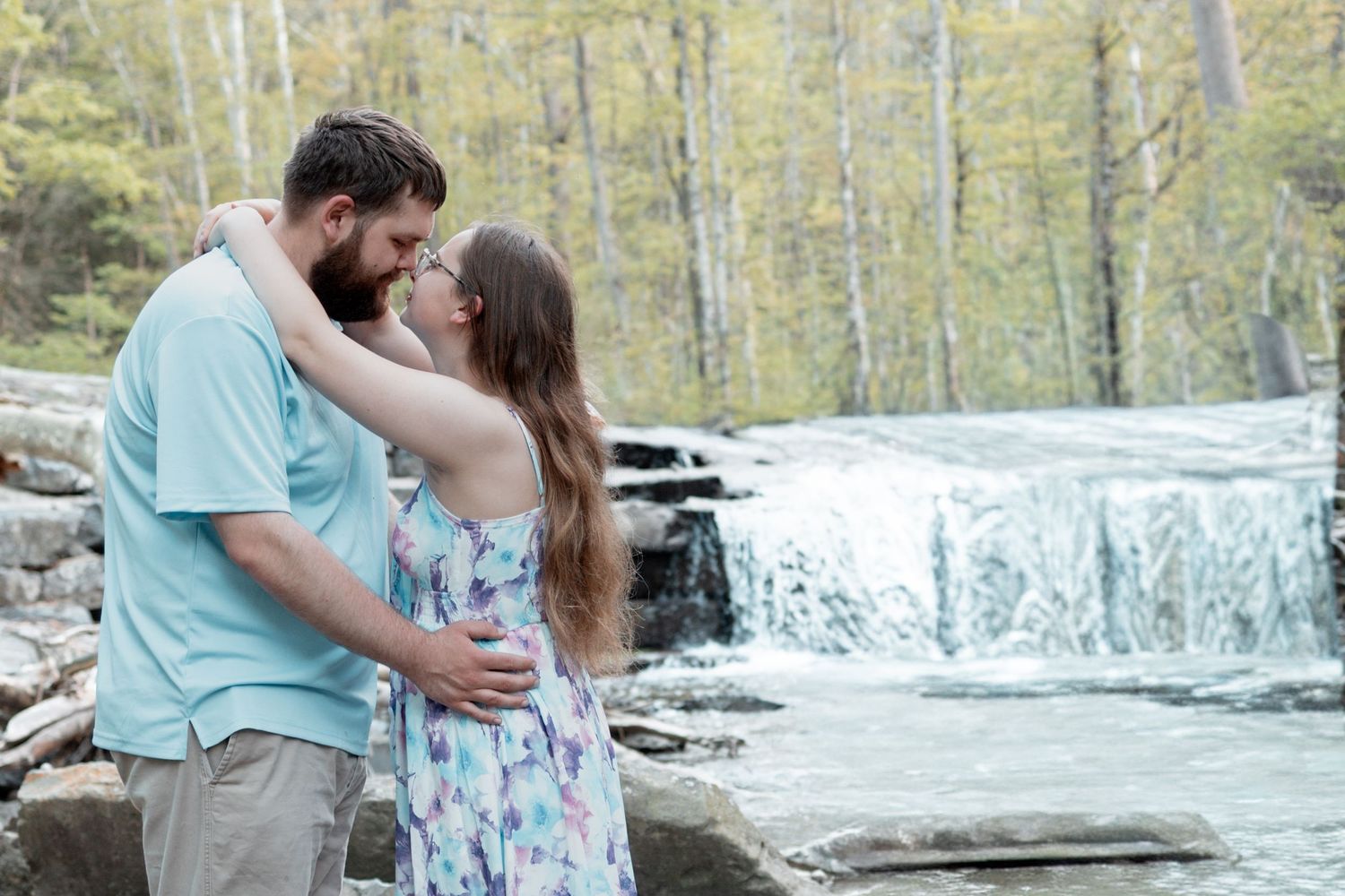 Couple embracing romantically in front of a scenic waterfall surrounded by lush green trees in a natural outdoor setting.