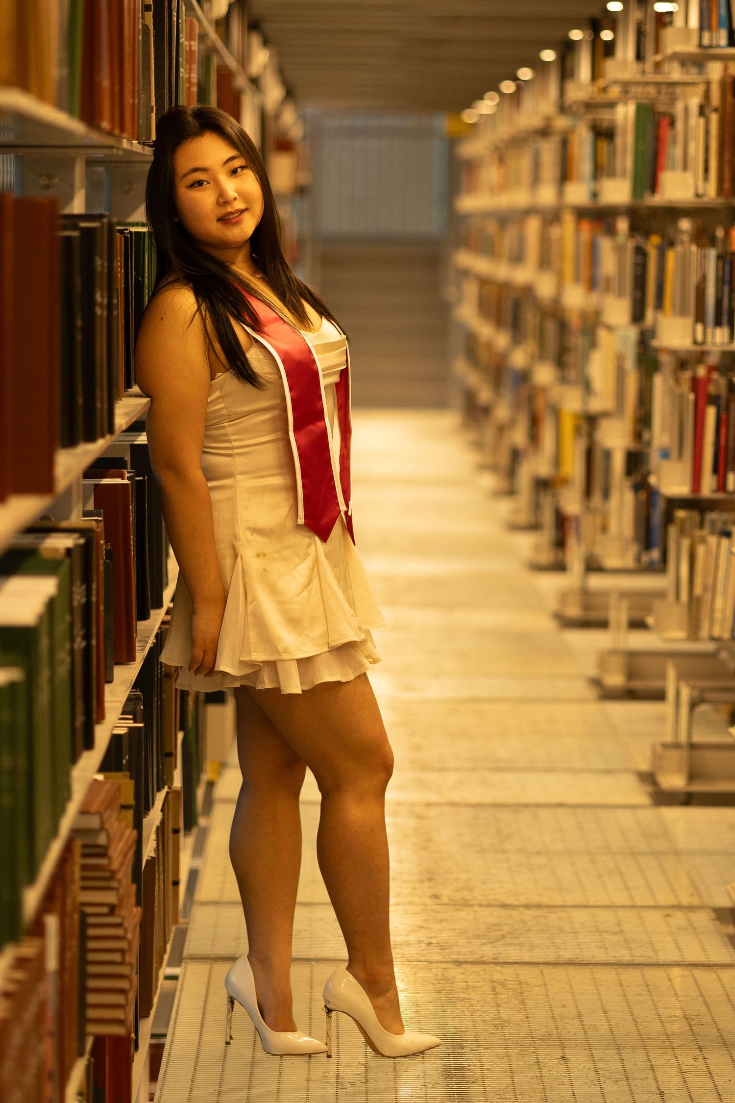 Young woman in white dress and red graduation sash poses in library aisle surrounded by bookshelves.