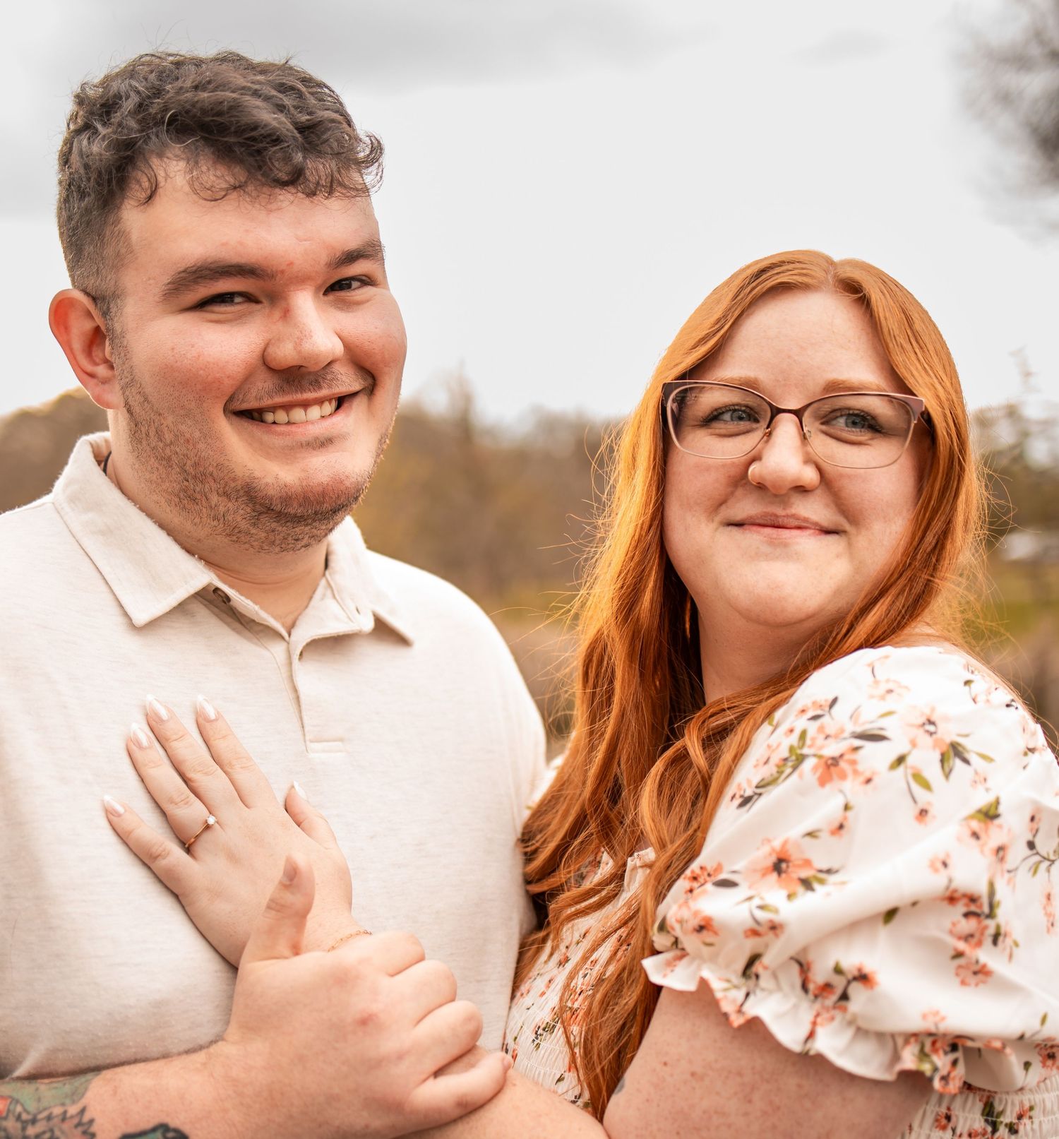 Smiling couple poses outdoors, woman with red hair and glasses in floral dress, man in beige polo shirt.