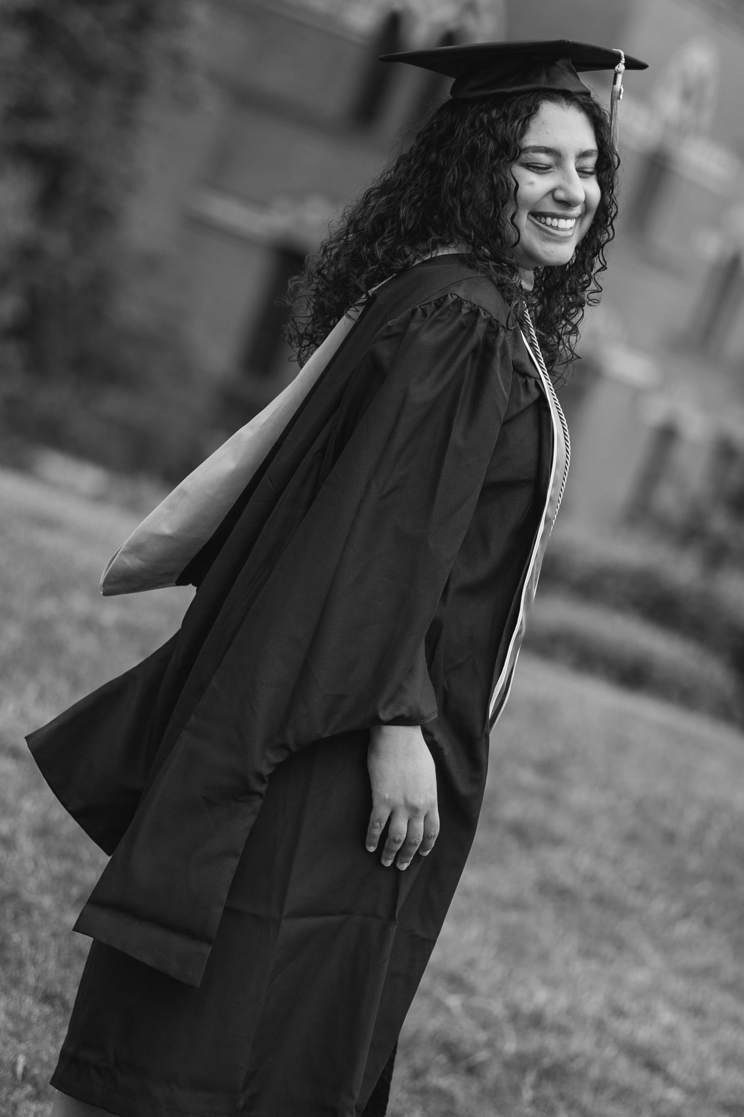 Black and white photo of a smiling young woman in graduation cap and gown, celebrating her academic achievement outdoors.