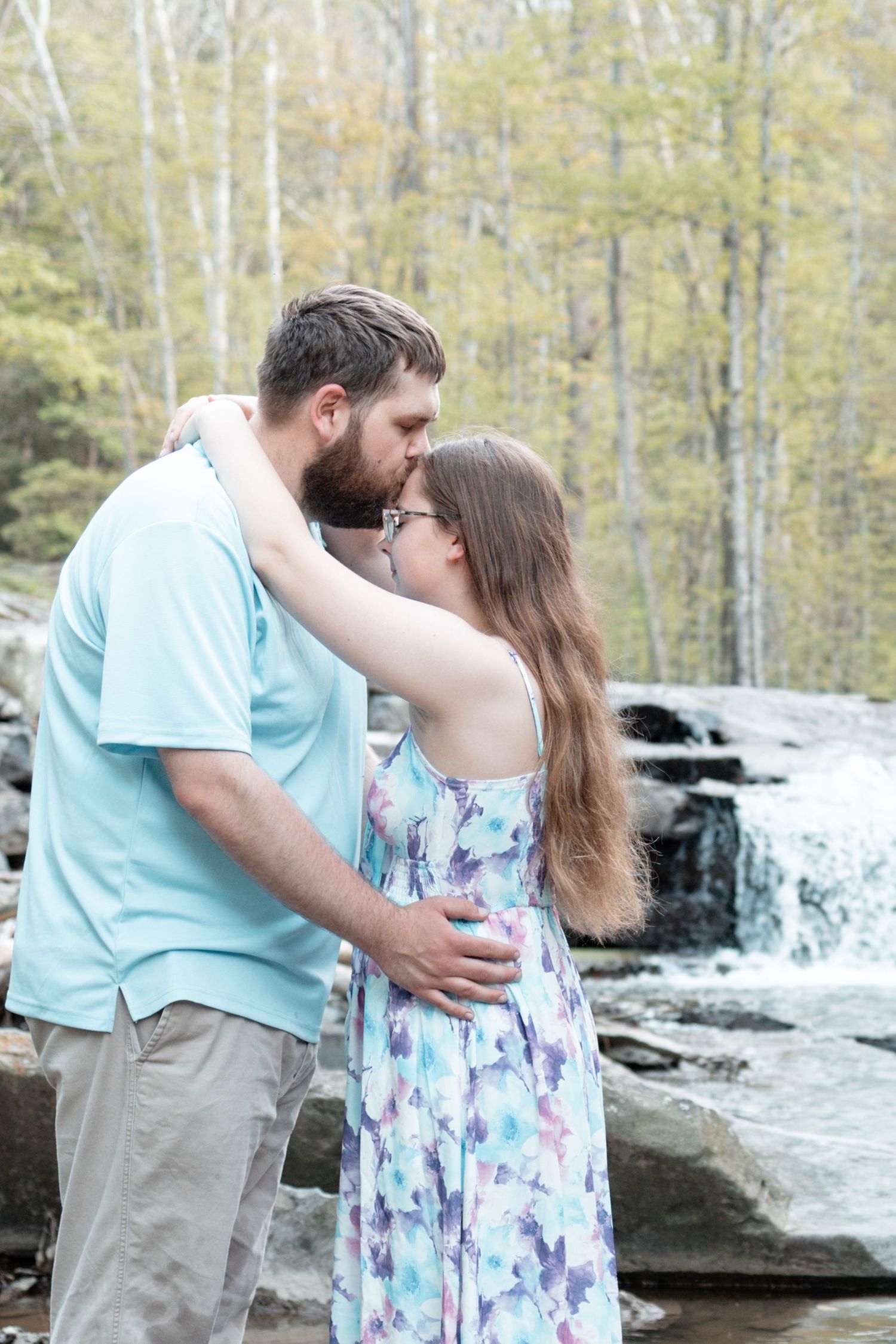 Couple sharing a kiss in front of a waterfall, woman in floral dress and man in light blue shirt embracing outdoors.