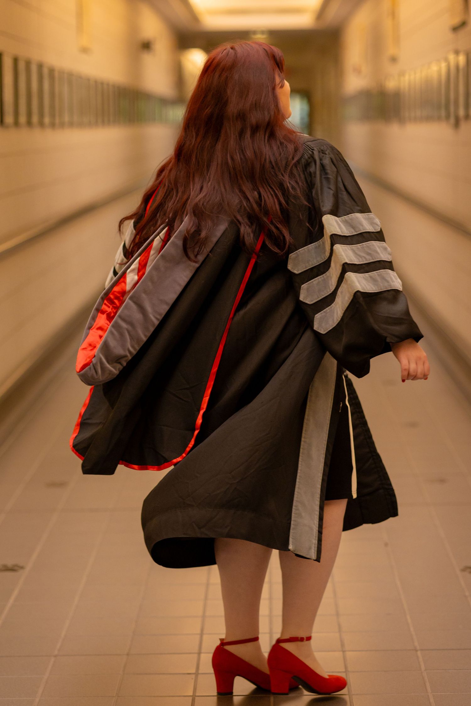 Woman in black graduation gown with red hood and red heels walks down a hallway, viewed from behind.