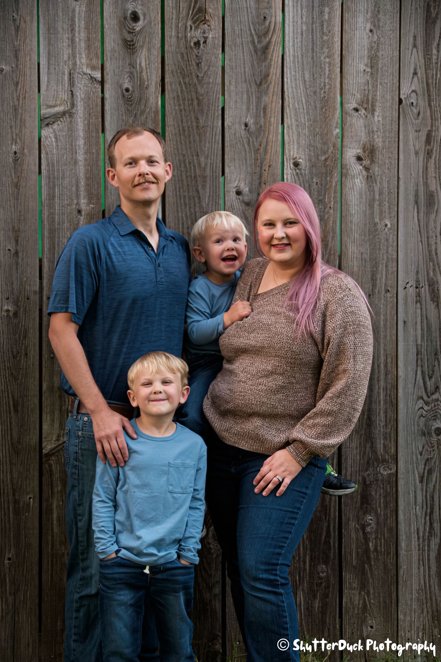 Family of four with two young boys posing together in front of a rustic wooden fence.