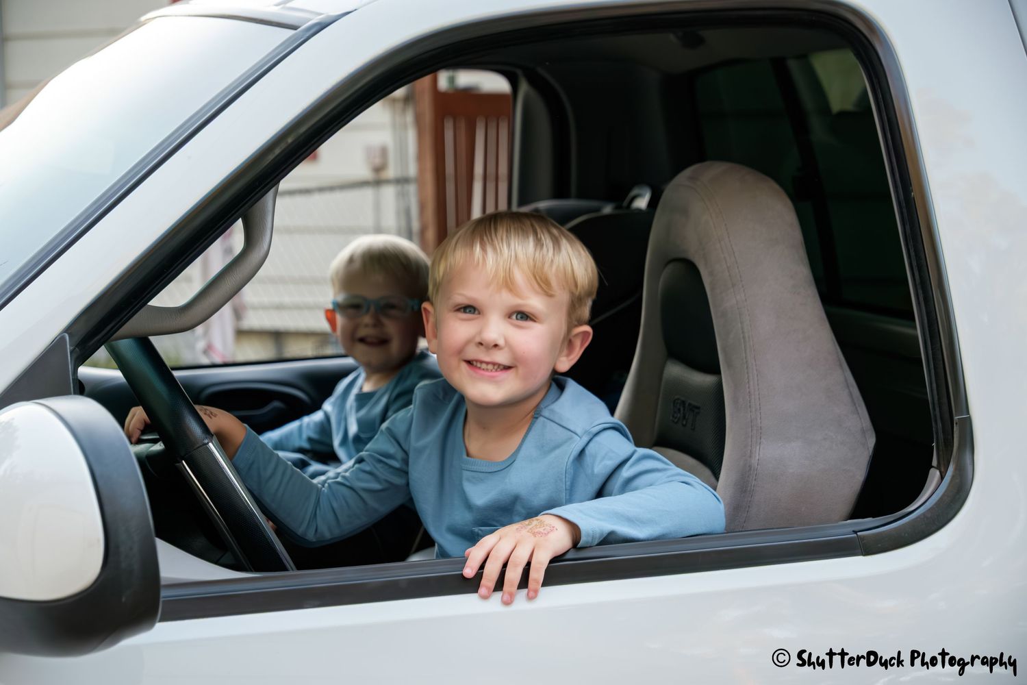 Two young blonde boys sitting in the open window of a white truck cab, smiling at the camera.