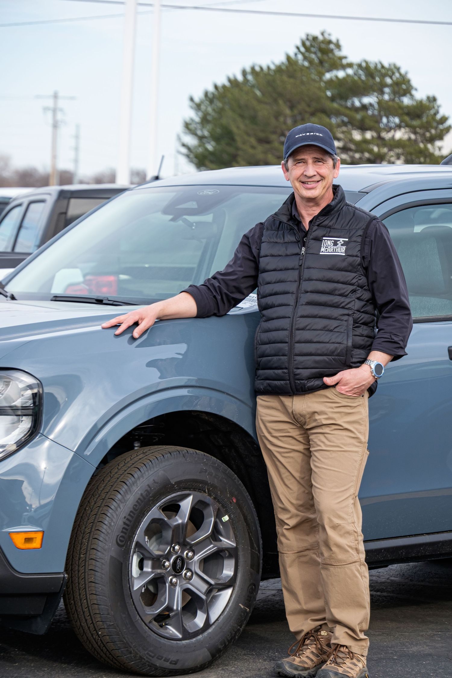 A smiling man in a black vest and cap leans against a blue Ford Maverick in a dealership lot.