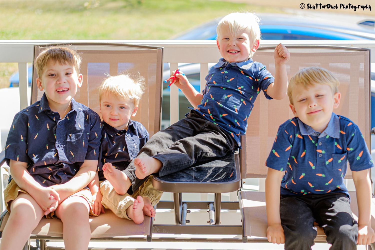 Four young boys in matching navy patterned shirts posing playfully on outdoor chairs.