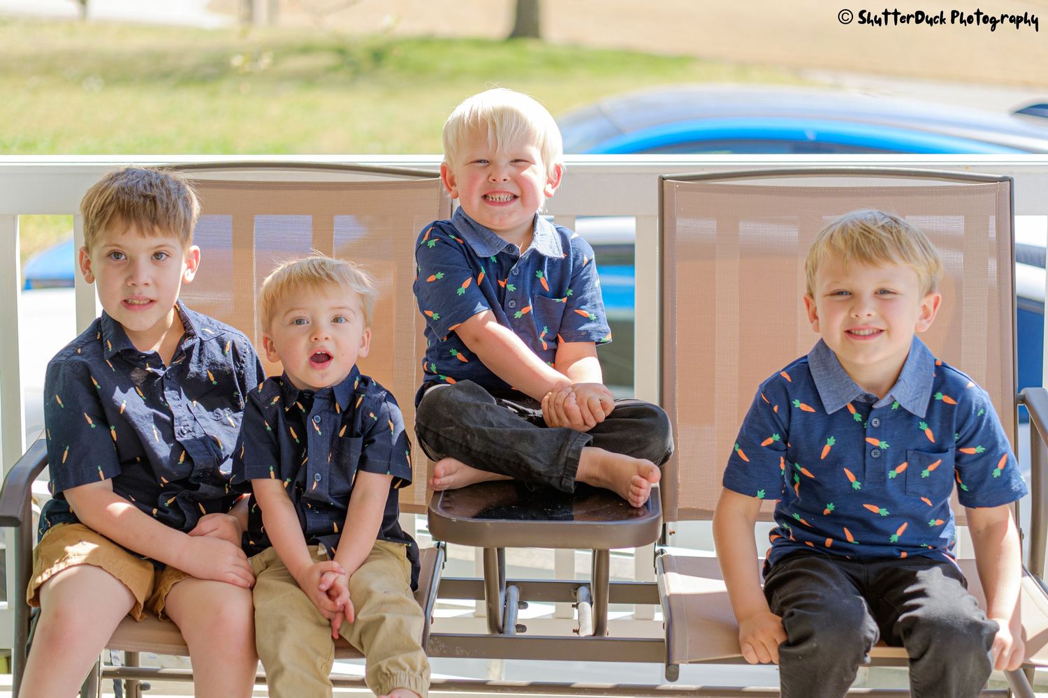 Four young blonde boys in matching navy shirts sitting together on outdoor lounge chairs smiling.