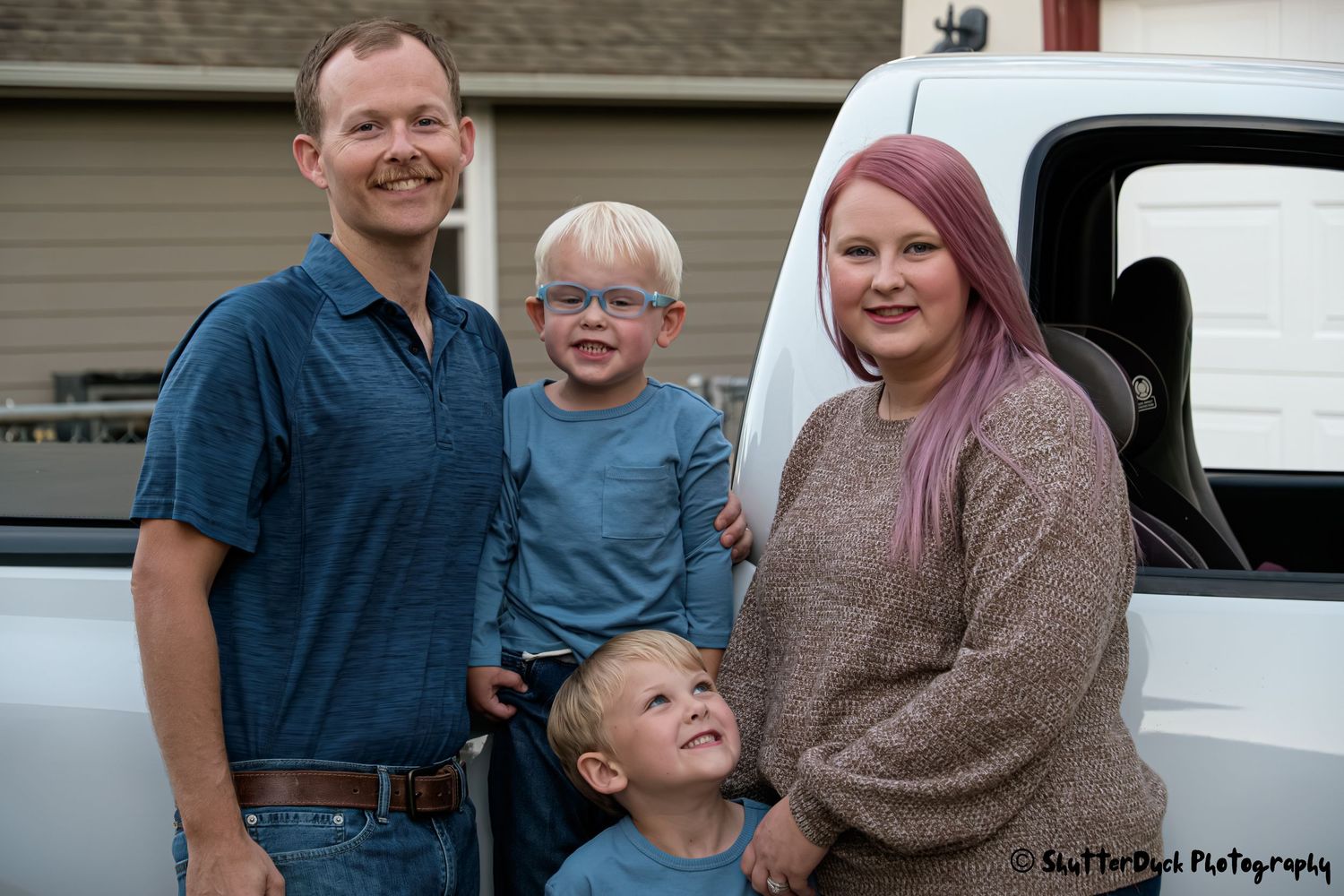 Happy family of four with two young boys posing beside a white pickup truck outdoors.