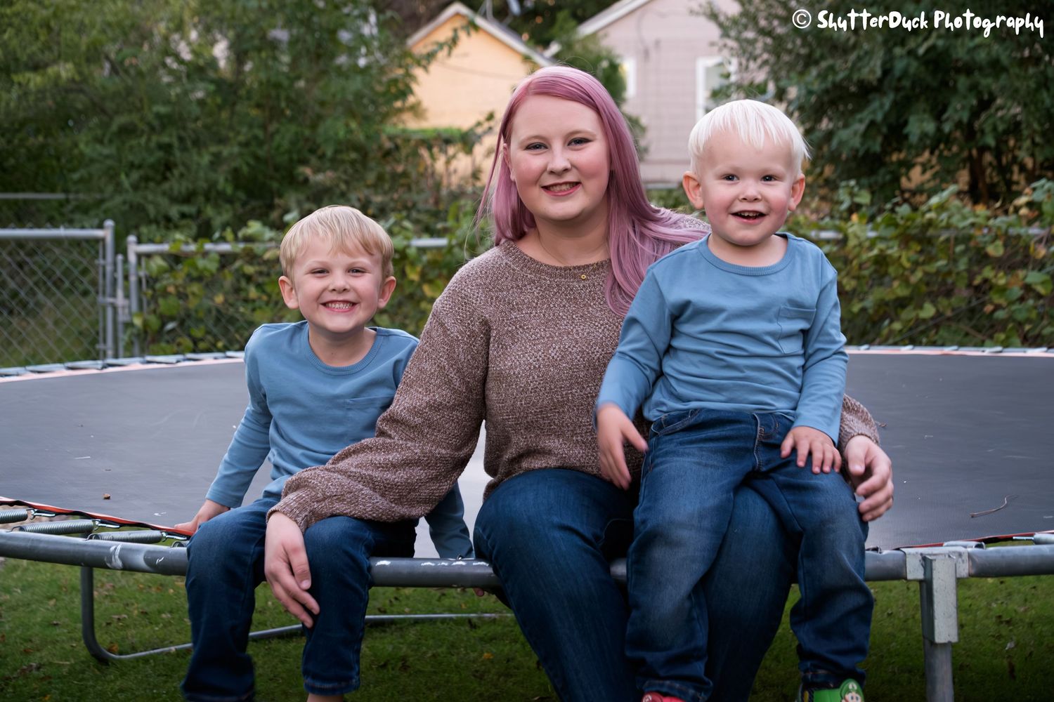 Smiling mother with pink hair sitting on a trampoline with her two young sons outdoors.