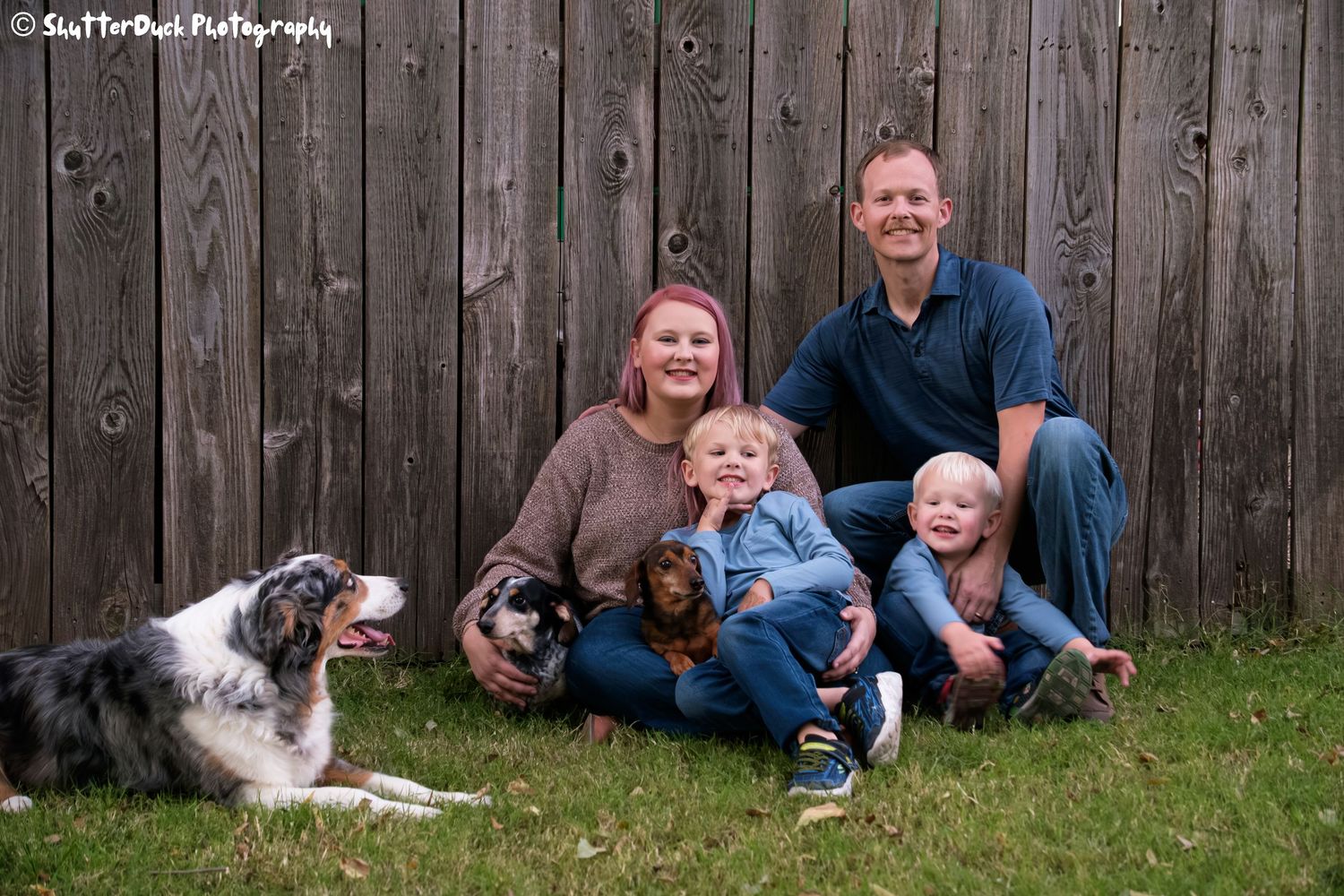 Happy family with two young children and three dogs sitting together in front of a wooden fence outdoors.
