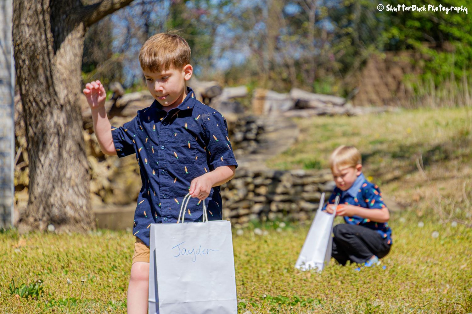 Two young boys collecting Easter eggs in gift bags on a sunny green lawn outdoors.