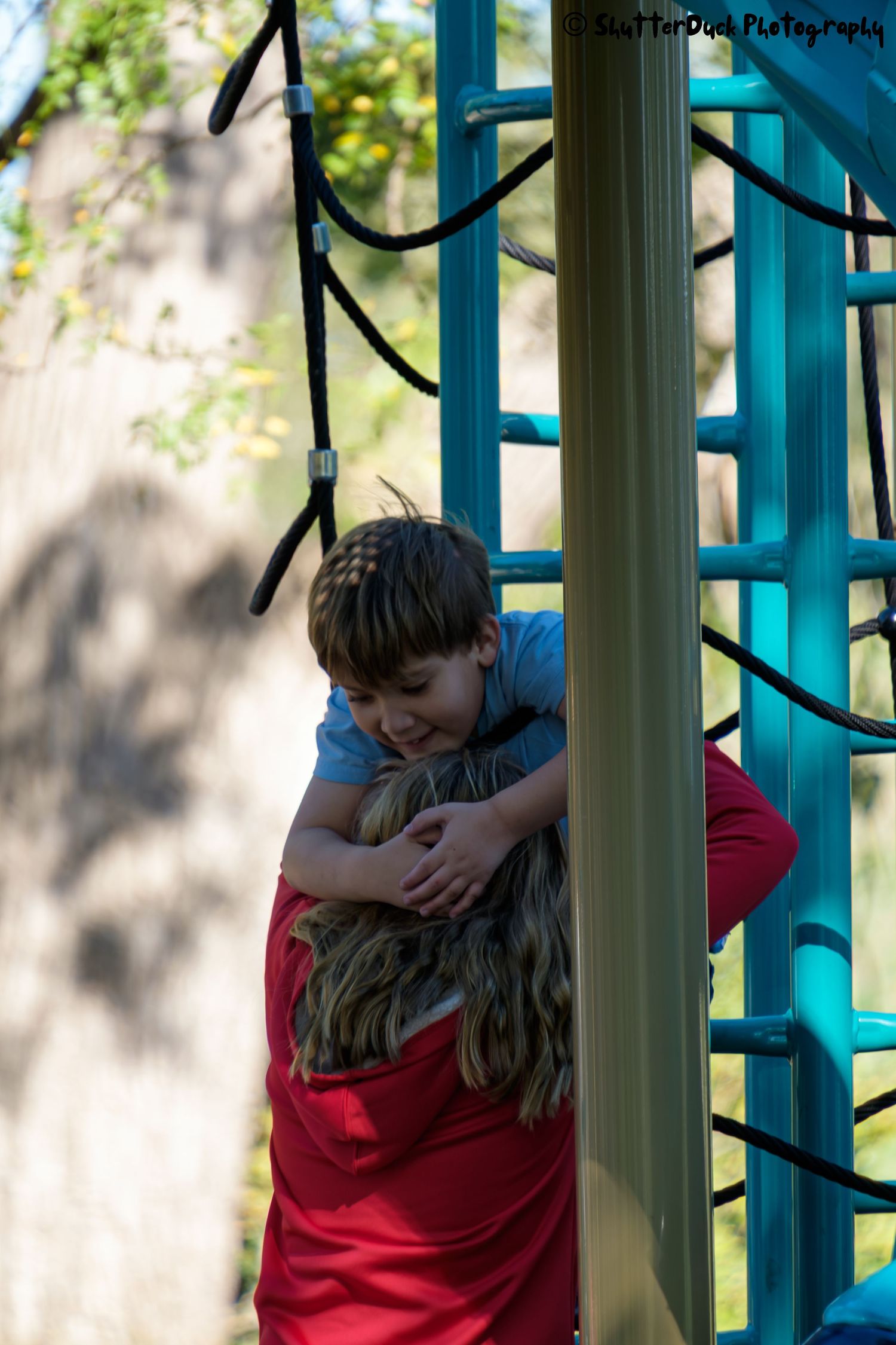 Two children share a sweet hug on a teal blue outdoor playground climbing structure.