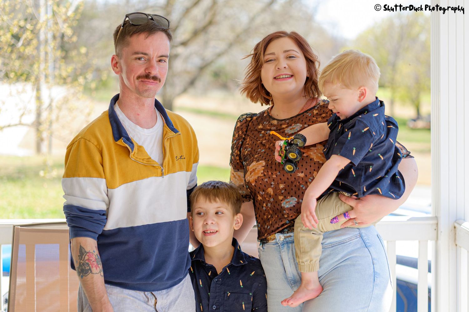 Young family of four with tattooed dad, mom holding toddler, and young son posing on a porch.