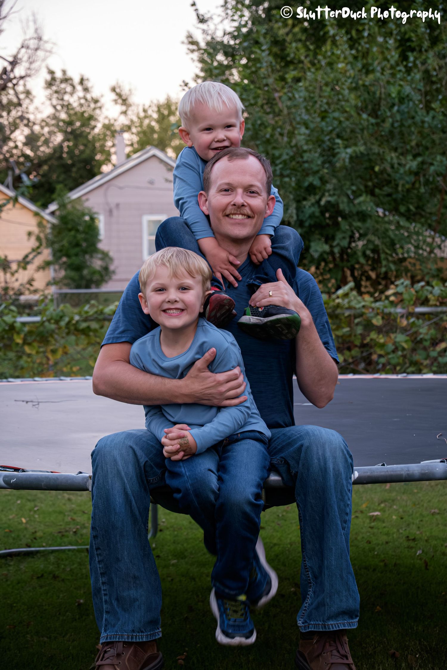 Father smiling with two young blonde boys stacking on his shoulders in a backyard near a trampoline.