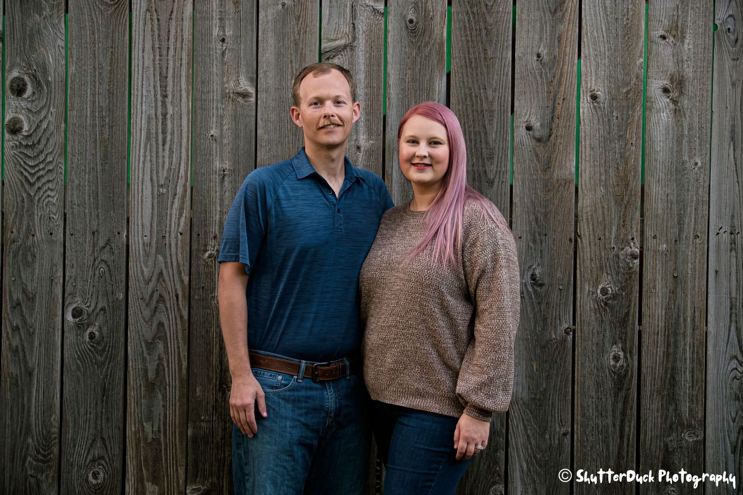 Smiling couple posing together in front of a rustic gray wooden fence outdoors.