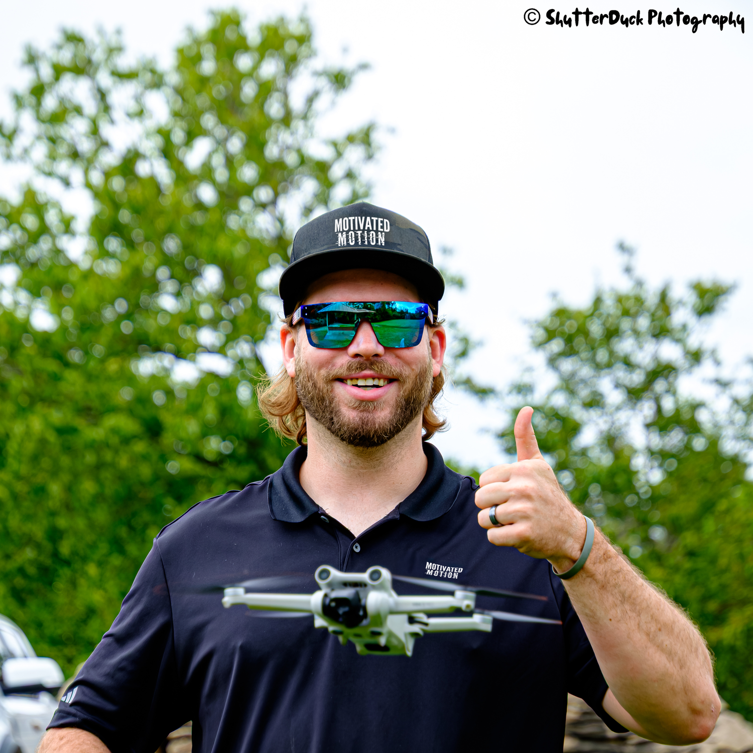Smiling man with beard giving thumbs up with hovering drone in front of him wearing Motivated Motion hat and sunglasses outdoors.