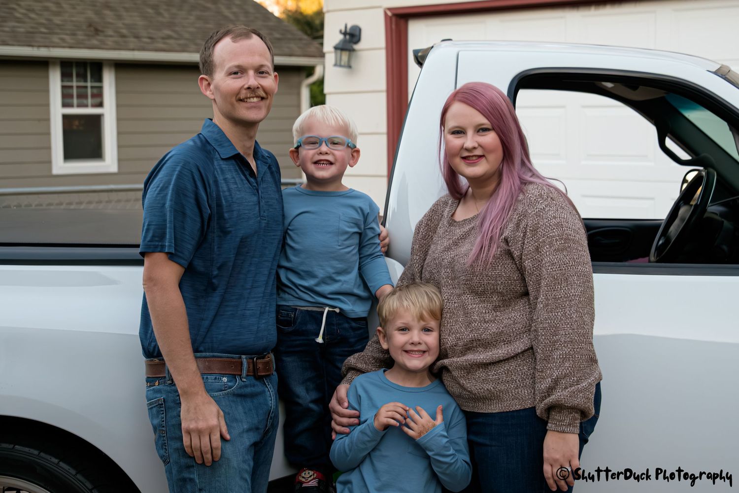 Family of four smiling together in front of a white pickup truck in a residential driveway at dusk.