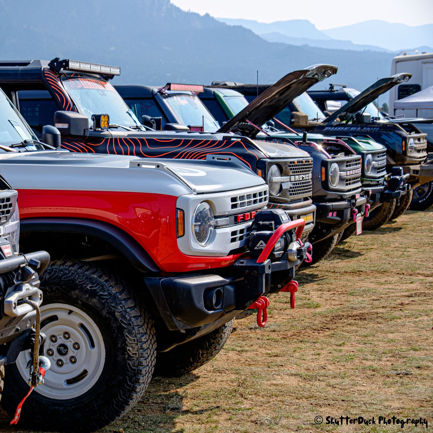 Line of rugged Ford Bronco 4x4 vehicles with winches at an outdoor off-road car show with mountains.