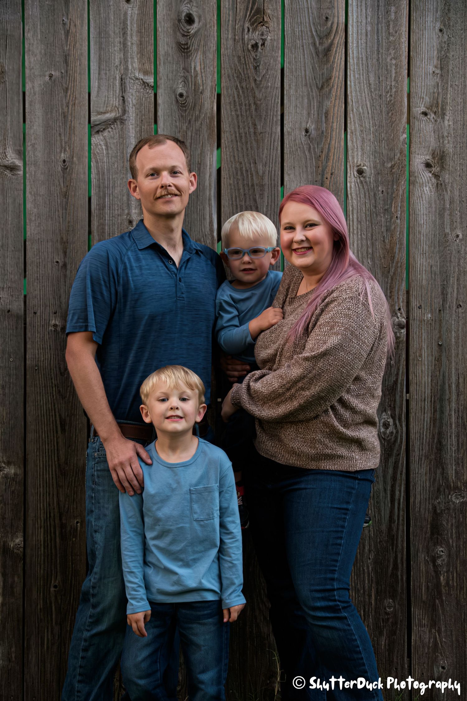 Family of four posing in front of a rustic wooden fence, parents with two young blonde boys.