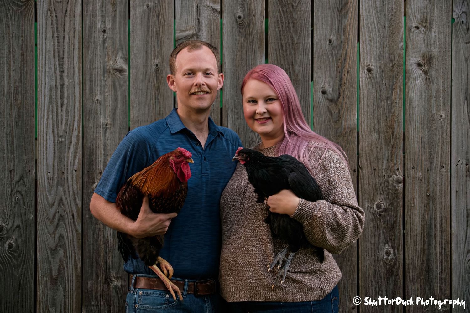 Smiling couple posing with a rooster and hen in front of a rustic wooden fence.