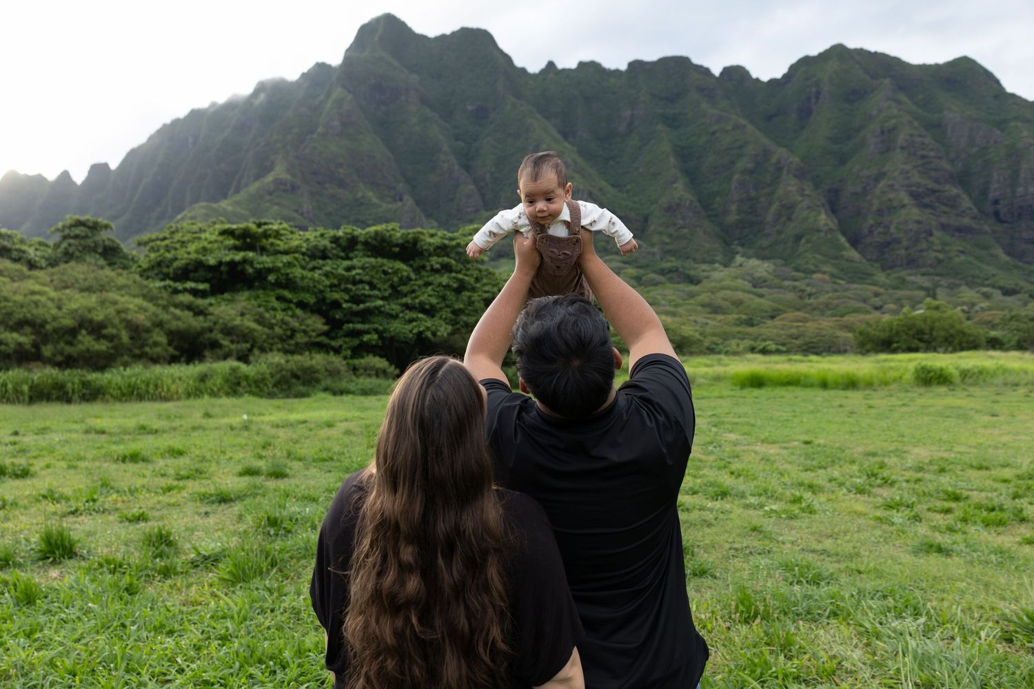 Parents lifting baby in air at Kualoa Ranch with dramatic green mountains in background Hawaii.